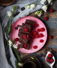 Load image into Gallery viewer, A selection of 6 Gluten-free, Dairy-free, Keto Raspberry brownies, on a pink plate with fresh raspberries. Set on a blue table surrounded by white lisanthus flowers on a light blue table cloth, with a cup of coffee to the side.
