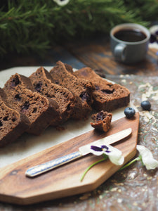 Nine Tea Cups Gluten Free Dairy Free Spiced Blueberry Breakfast Bread Slices on a table, next to sprigs of pine, a cup of coffee, white and purple pansies and a blue enamel bread knife