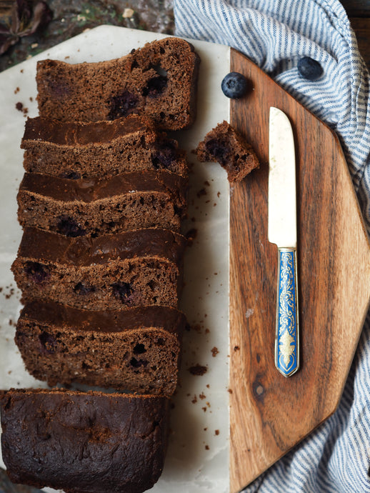 Nine Tea Cups Gluten Free Dairy Free Spiced Blueberry Breakfast Bread Sliced on an octagonal marble chopping board, with fresh blueberries, placed on a striped cloth and next to a blue enamel bread knife