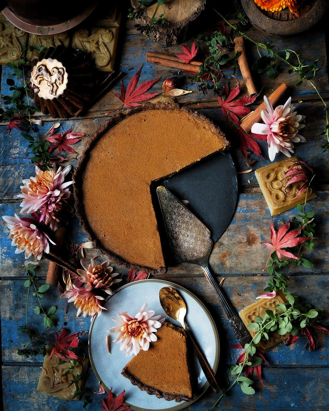 Nine Tea Cups Bakery Raw Vegan, Gluten Free, Dairy Free Pumpkin Pie on a black plate surrounded by flowers, autumn leaves, and beeswax candles, with a slice taken out and placed on a grey plate next to it with a gold coloured spoon and a peach-copper flower